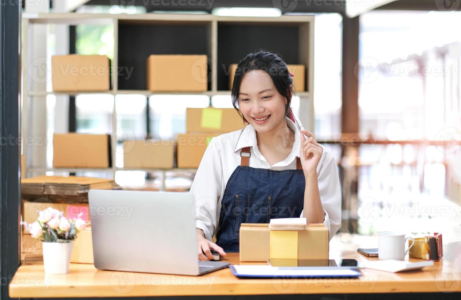 Start-up-KMU-Kleinunternehmer einer freiberuflichen Asiatin mit Schürze, die mit Laptop und Box Bestellungen online entgegennimmt und überprüft, um sich auf den Verkauf an Kunden vorzubereiten, Online-KMU-Geschäftsideen. foto