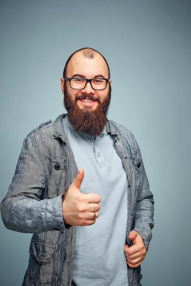 Der Lebensstil eines erfolgreichen jungen Mannes mit Brille, Bart, modischer Jeansjacke mit Daumen nach oben, emotionales Männerporträt im Studio foto