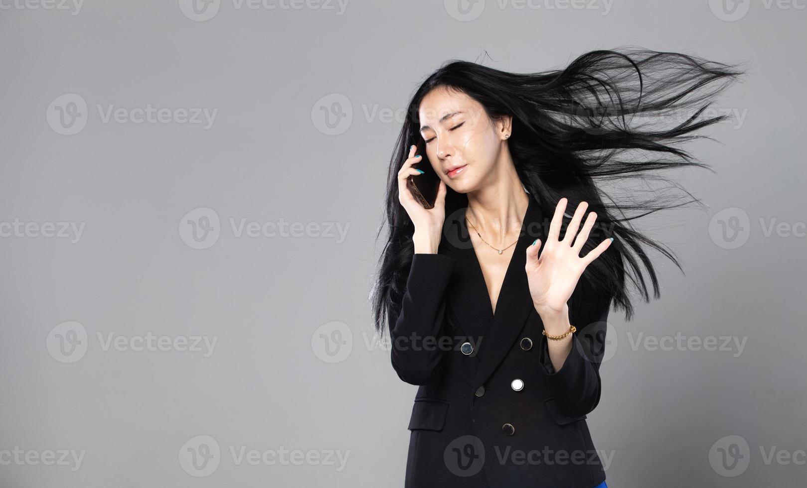 asiatische transgender-frau mit langen schwarzen glatten haaren, windschlag in die luft werfen. weibliches grifftelefon gegen windsturm, gefühl mode sinnlich sexy, grauer hintergrund isoliert kopienraum foto