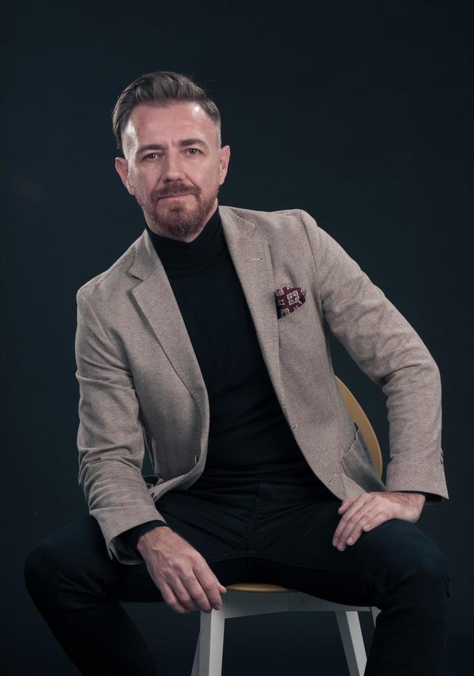 Porträt eines erwachsenen Geschäftsmannes, der einen trendigen Anzug trägt und in einem modernen Studio auf einem stilvollen Stuhl vor dem schwarzen Hintergrund sitzt foto