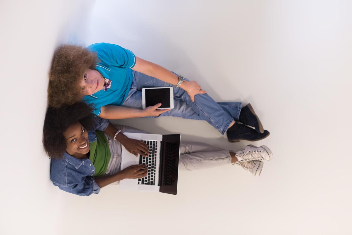 Multiethnisches Paar, das mit einem Laptop und einer Tablet-Draufsicht auf dem Boden sitzt foto