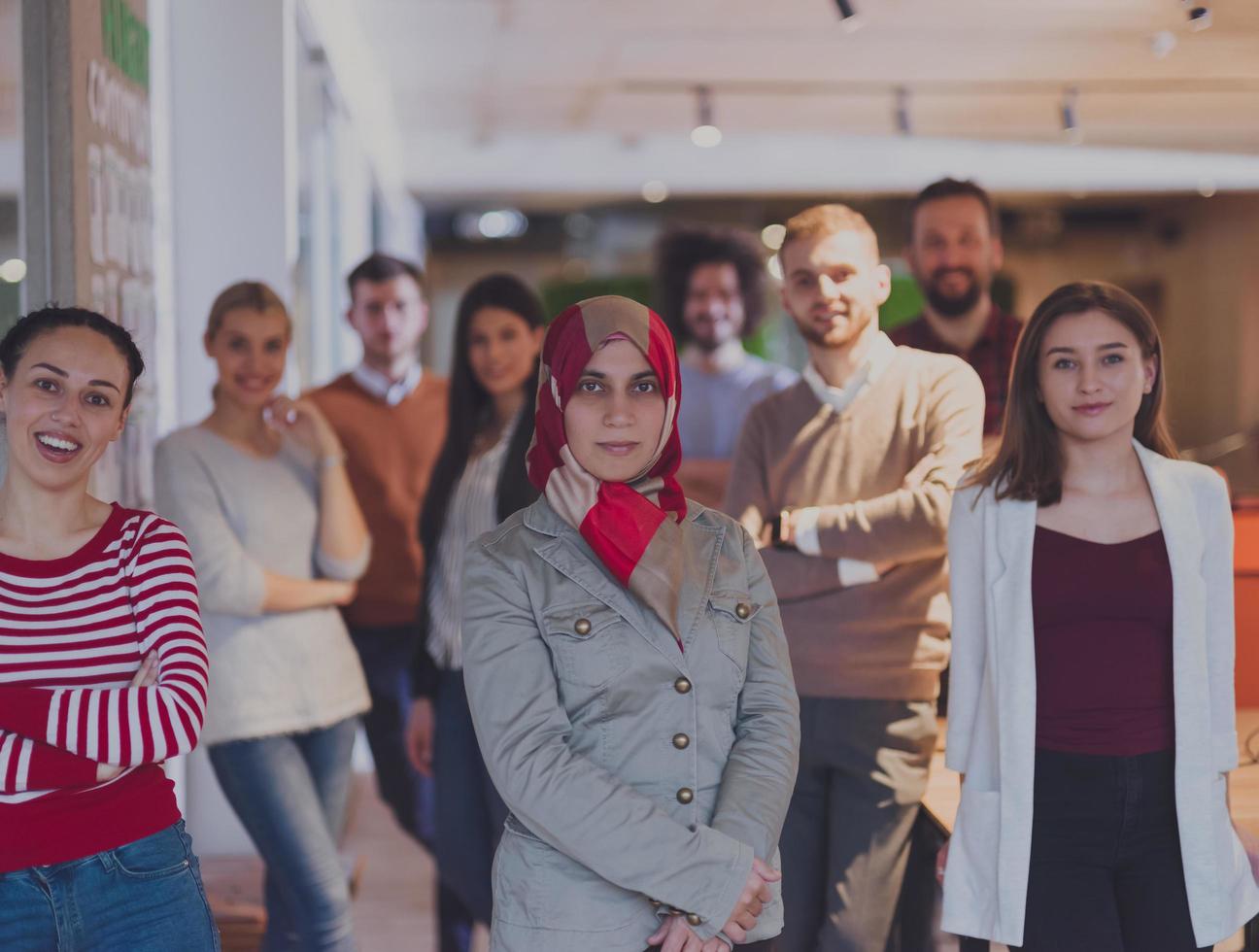 schöne arabische geschäftsfrau im hijab, die in die kamera schaut und lächelnde kollegen vielfältiges team in einem modernen open space startup office im hintergrund foto