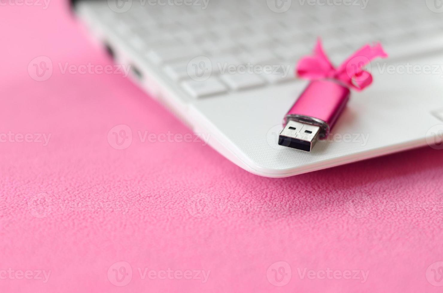 Eine leuchtend rosa USB-Flash-Speicherkarte mit einer rosa Schleife liegt auf einer Decke aus weichem und pelzigem hellrosa Fleece-Stoff neben einem weißen Laptop. klassisches weibliches Geschenkdesign für eine Speicherkarte foto