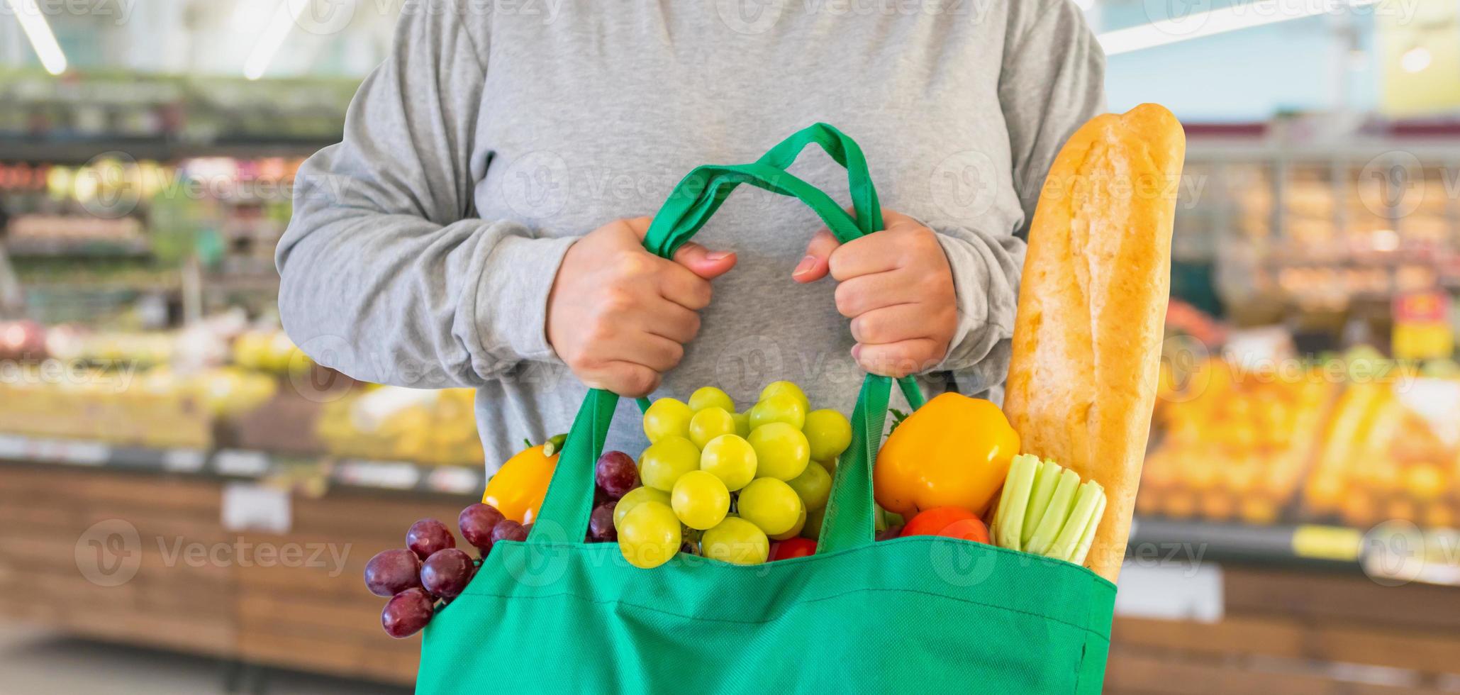 Kunden halten wiederverwendbare grüne Einkaufstasche mit Obst und Gemüse über Supermarktgang verschwimmen defokussierte Produktregale im Inneren von Bokeh hellem Hintergrund foto