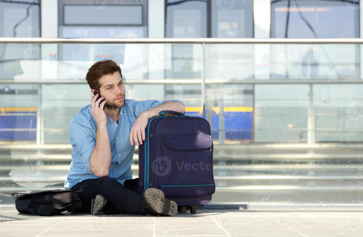 männlicher Reisender, der auf Boden sitzt und auf Handy spricht foto