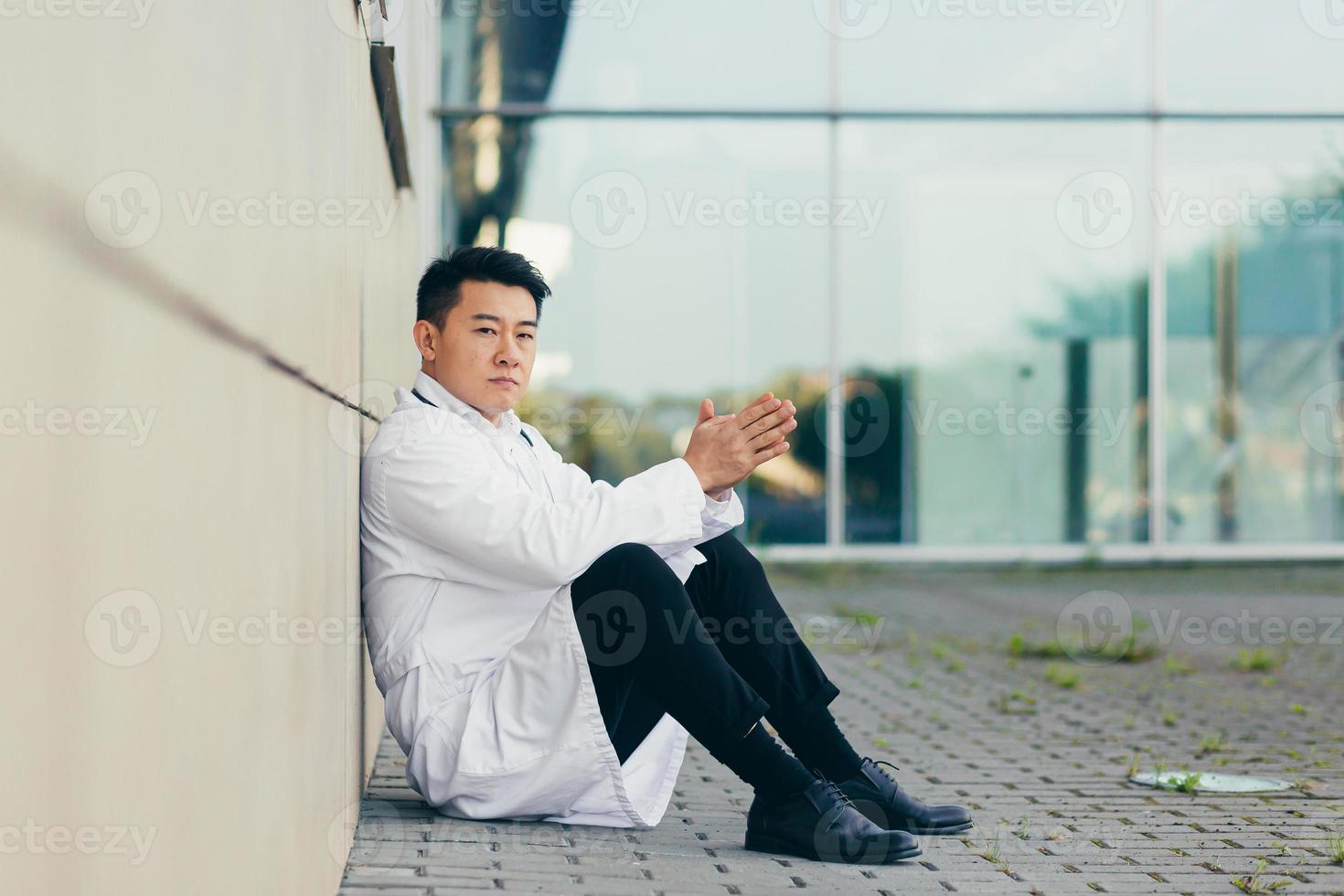 porträt arzt asiatischer mann müde nach der arbeit auf dem boden in der nähe der klinik sitzend enttäuscht foto