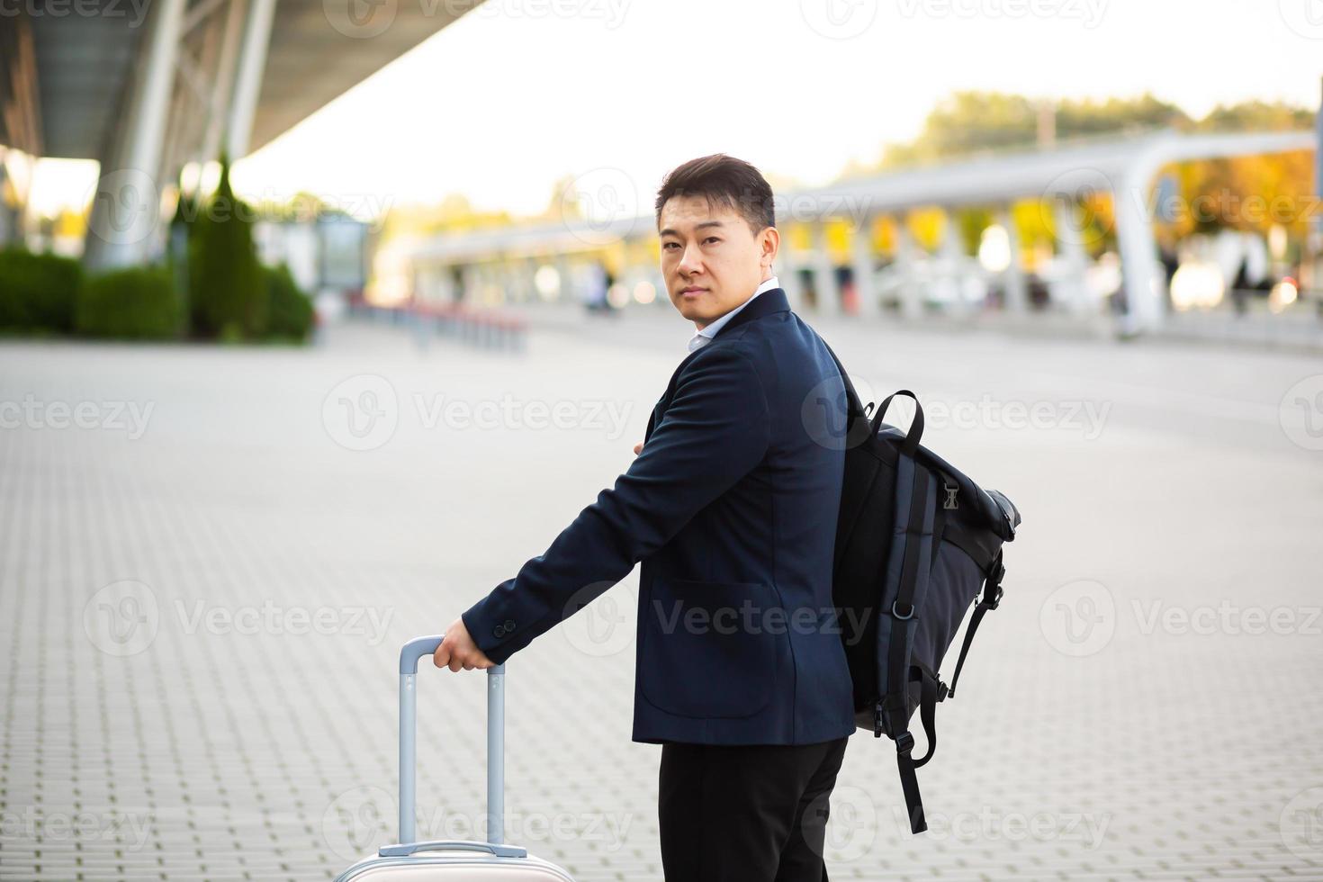 erfolgreicher asiatischer geschäftsmann in der nähe von flughafen und busbahnhof geht mit koffern ernst foto