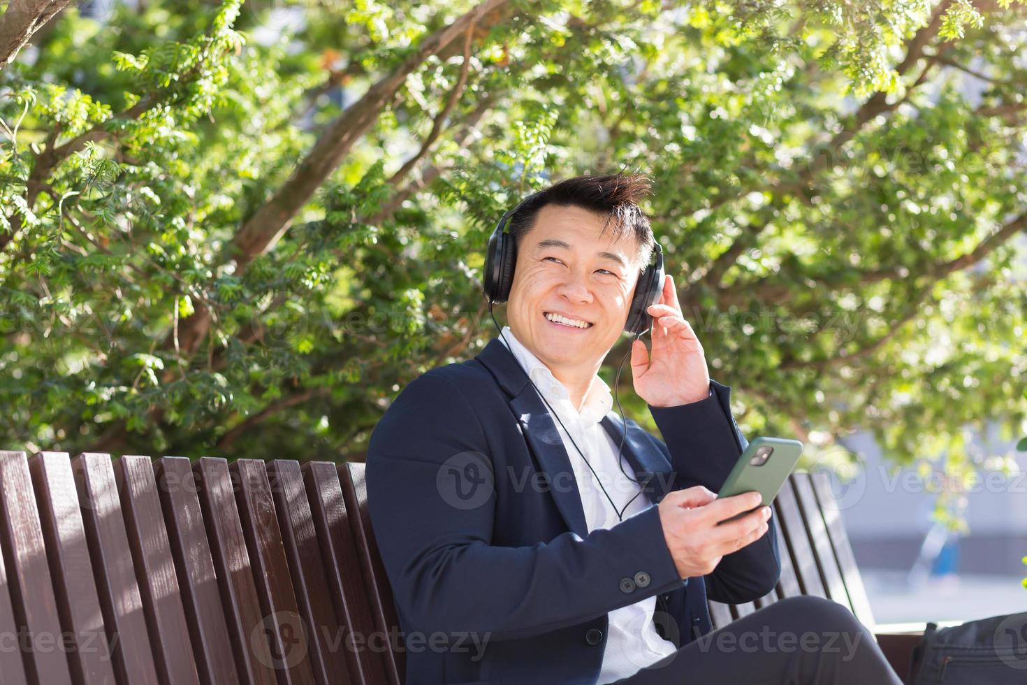 Fröhlicher asiatischer Geschäftsmann, der auf einer Bank in einem Stadtpark in der Innenstadt sitzt und Musik hört foto