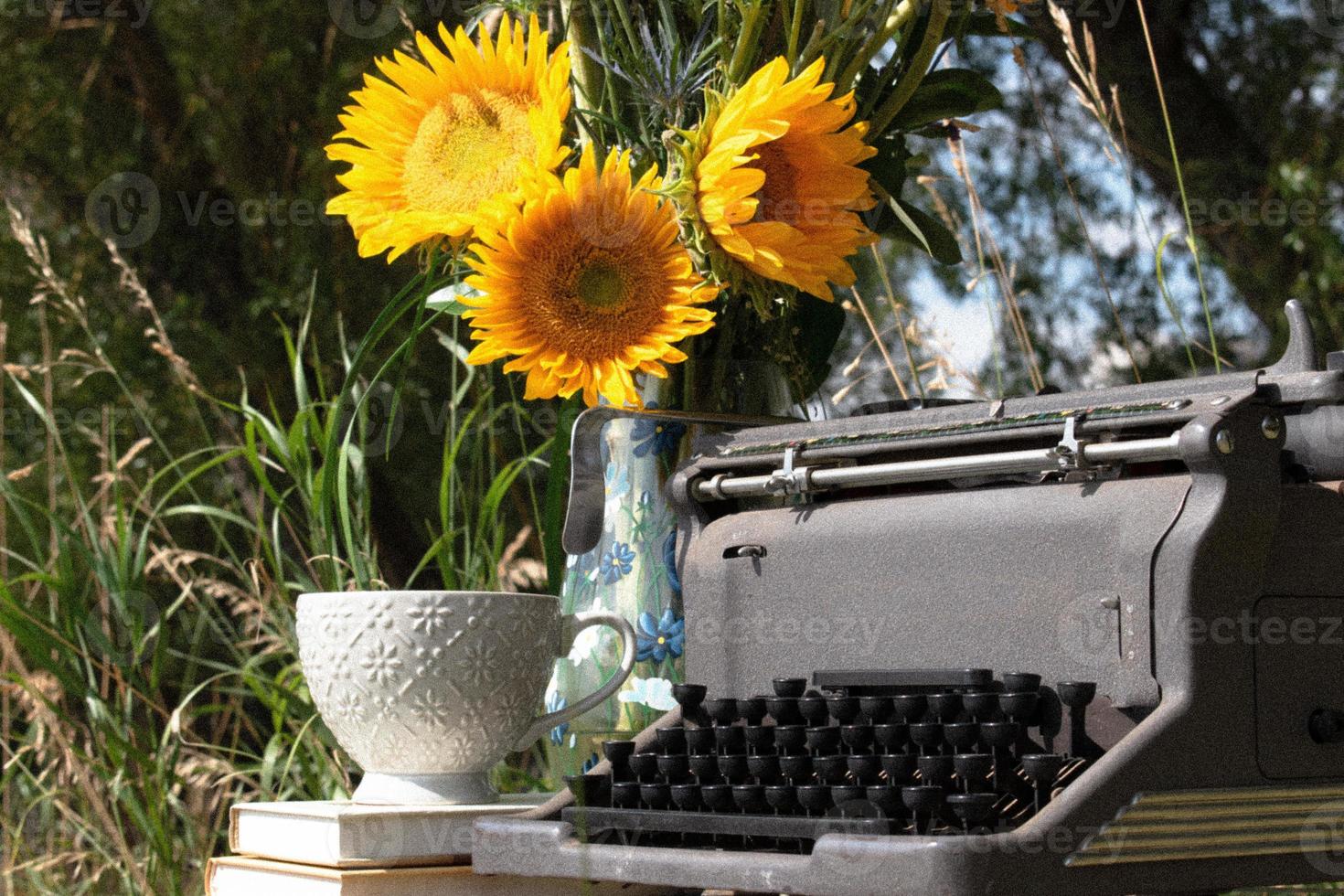 Vintage-Schreibmaschine unter einem Baum mit Sonnenblumen, Büchern und einer Teetasse foto