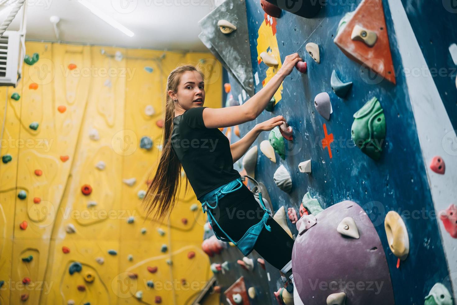 junge Frau, die eine hohe, künstliche Kletterwand im Innenbereich erklimmt foto