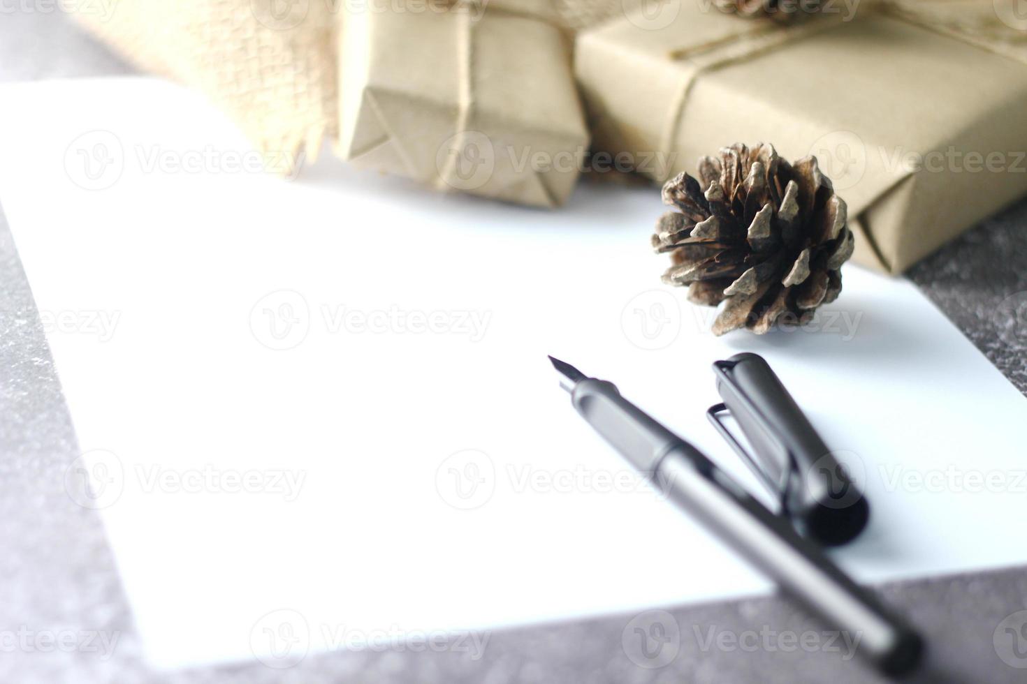 feiertagsgeschenke weihnachts- und neujahrsgeschenkbox in braunem papier, tannenzapfen und weißem papier und stiften auf dem tisch platziert. foto