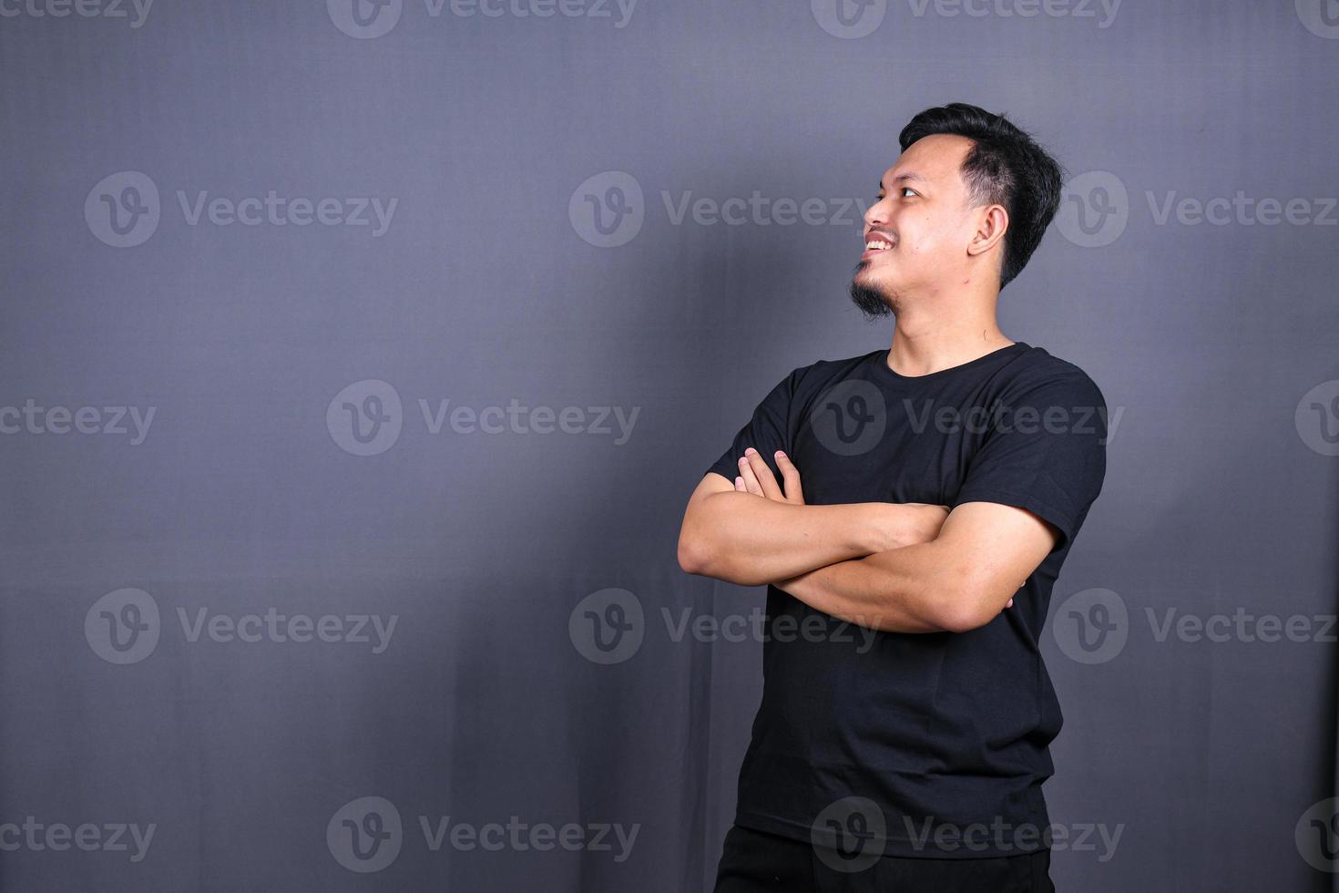 lächelnder gutaussehender asiatischer mann im schwarzen t-shirt, das mit verschränkten armen auf grauem hintergrund steht foto