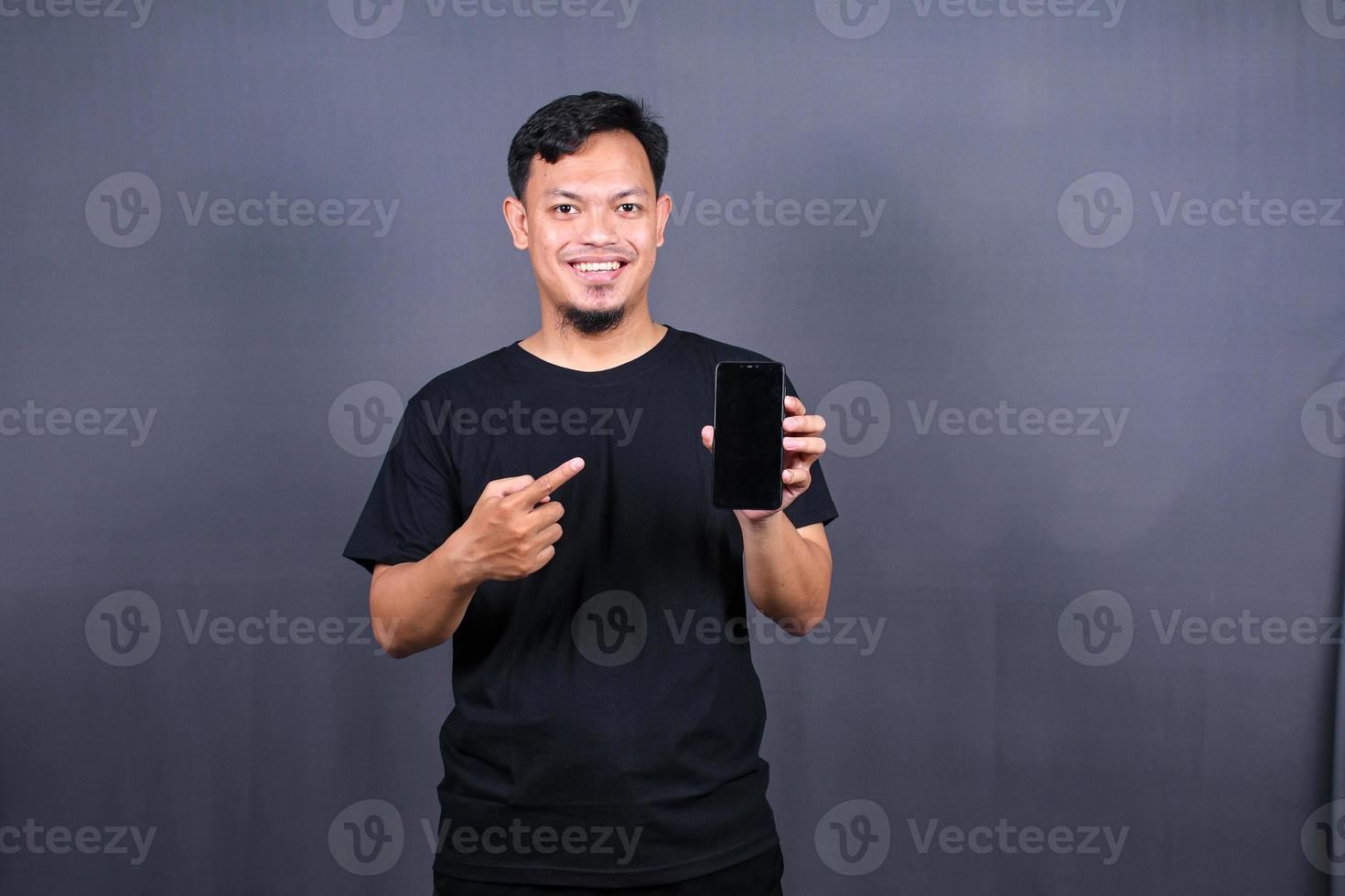 Porträt eines jungen asiatischen Mannes mit schwarzem T-Shirt, der isoliert vor grauem Hintergrund steht und mit dem Finger auf das Handy zeigt foto
