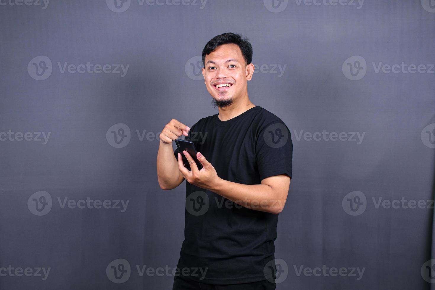 Porträt eines jungen asiatischen Mannes mit schwarzem T-Shirt, der isoliert vor grauem Hintergrund steht und mit dem Finger auf das Handy zeigt foto