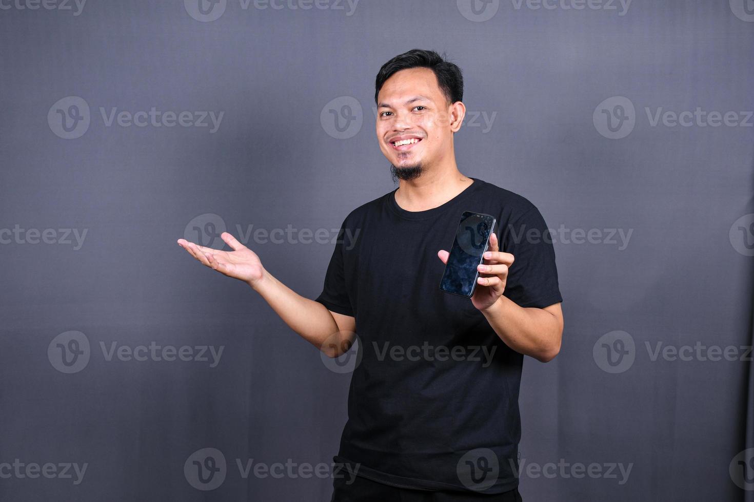 foto des attraktiven asiatischen kerls, der handy hält, trägt schwarzes t-shirt, isolierter grauer farbhintergrund