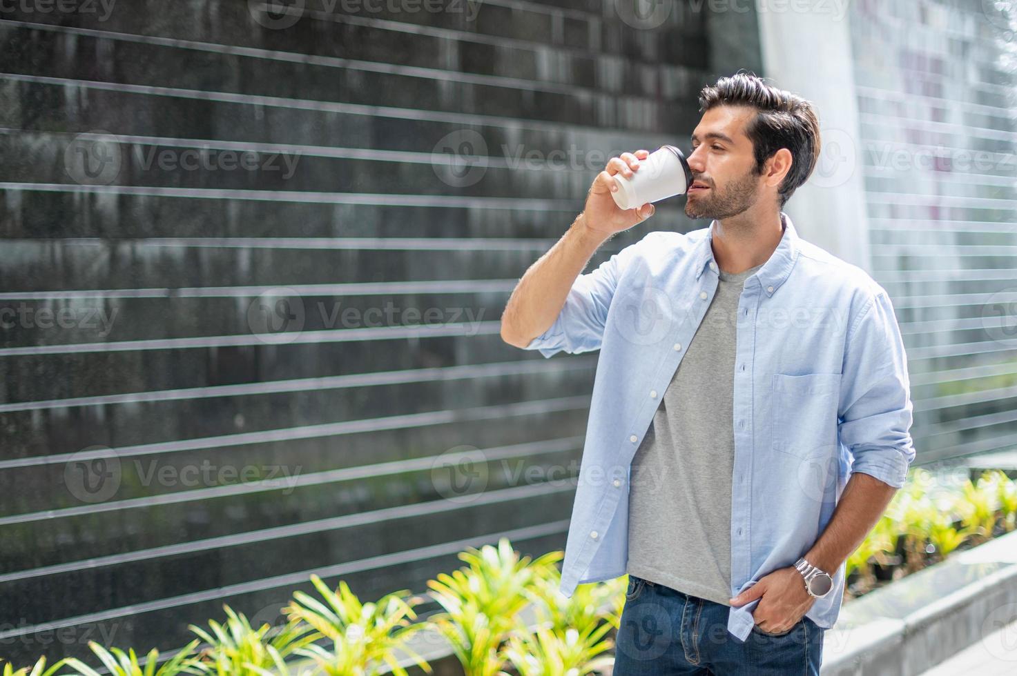 junger kaukasischer mann, der kaffee trinkt und ein lässiges hemd trägt, während er auf seinem weg geht, um sich in seinem urlaub zu entspannen. foto