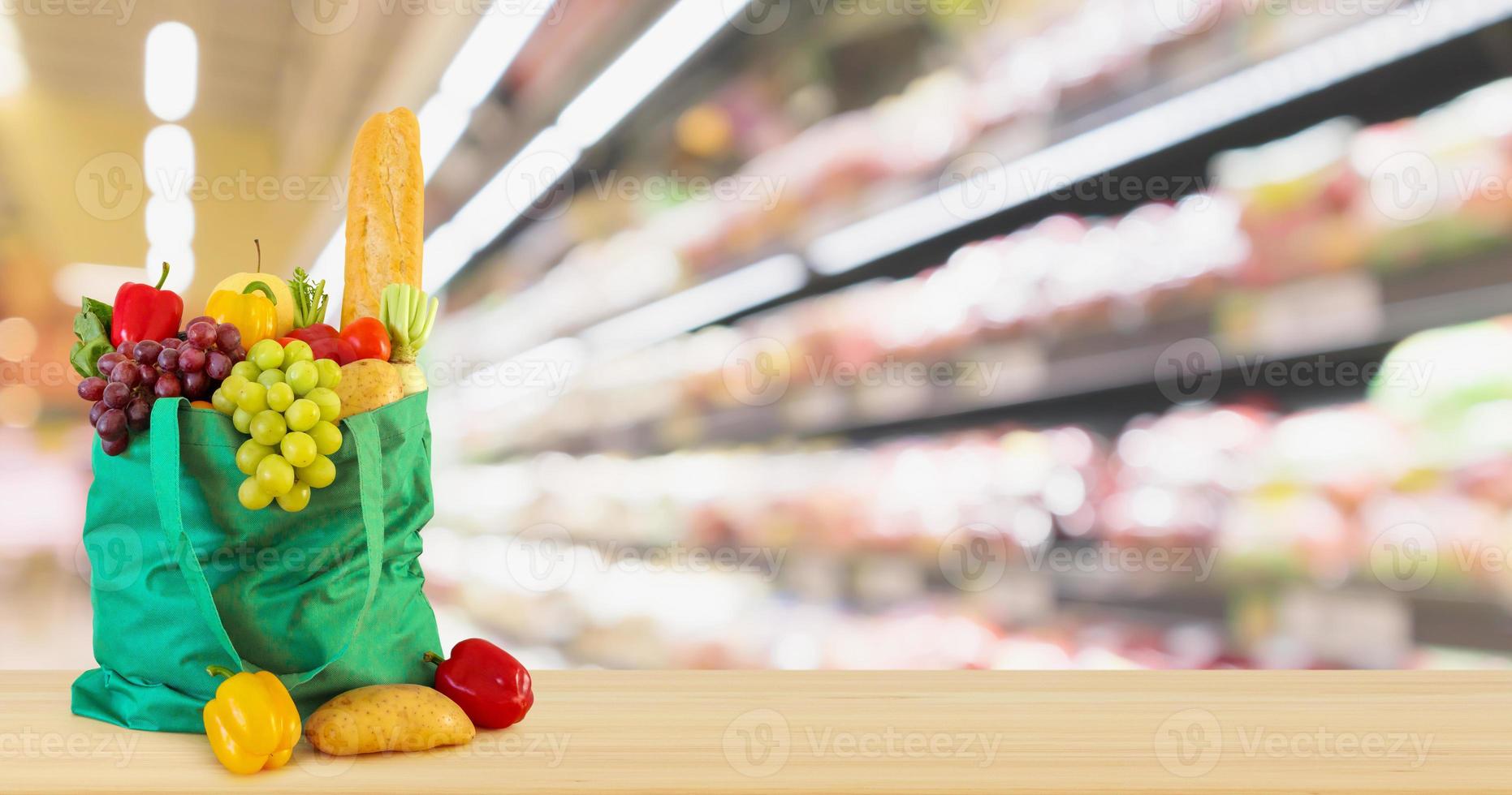 frisches obst und gemüse in wiederverwendbarer grüner einkaufstasche auf holztischplatte mit supermarkt-lebensmittelgeschäft unscharfer defokussierter hintergrund mit bokeh-licht foto