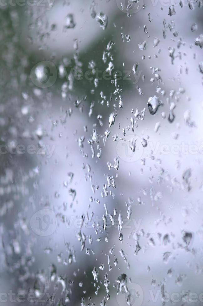 ein Foto von Regentropfen auf dem Fensterglas mit verschwommenem Blick auf die blühenden grünen Bäume. abstraktes Bild, das bewölkte und regnerische Wetterbedingungen zeigt