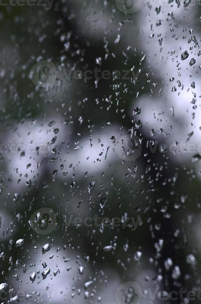 ein Foto von Regentropfen auf dem Fensterglas mit verschwommenem Blick auf die blühenden grünen Bäume. abstraktes Bild, das bewölkte und regnerische Wetterbedingungen zeigt