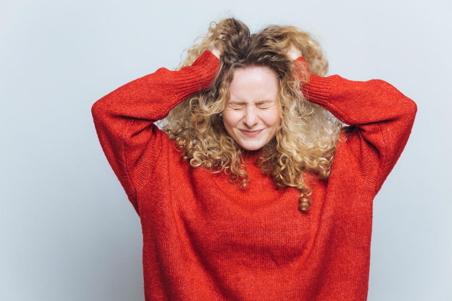 stressige frustrierte blonde frau reißt haare aus, bedauert fehler, drückt negative gefühle aus, trägt lockeren roten pullover, isoliert über weißem studiohintergrund. emotionale junge weibliche drinnen foto