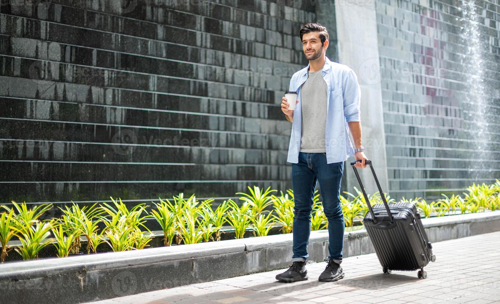 junger kaukasischer mann, der kaffee trinkt und den koffer zieht und auf seinen freund wartet, um gemeinsam zu reisen. foto