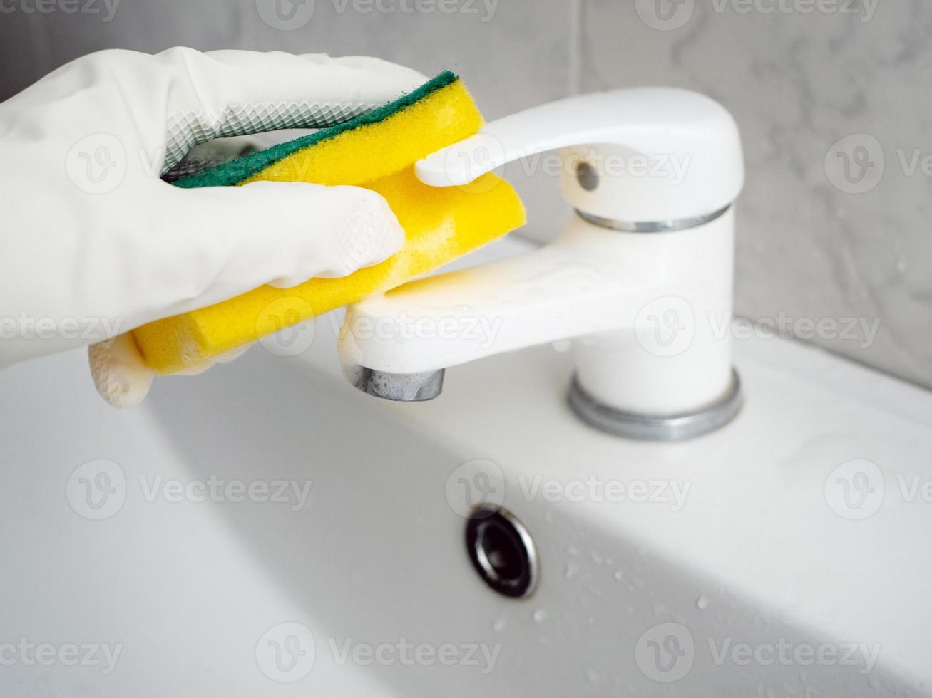 Eine Hand in einem weißen Gummihandschuh mit einem Schwamm wäscht einen weißen Wasserhahn im Badezimmer foto
