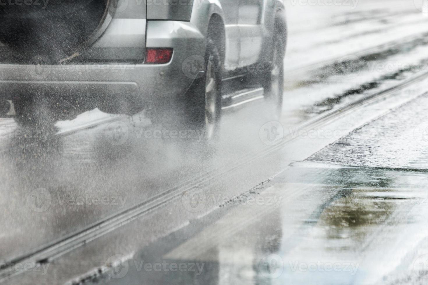 Regenwasserspritzer fließen von den Rädern des silbernen Crossover-Autos, das sich schnell in der Tageslichtstadt mit selektivem Fokus bewegt foto