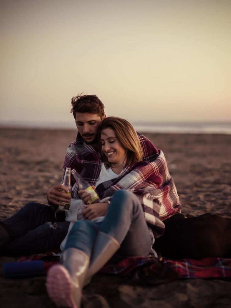 Liebendes junges Paar, das am Strand neben dem Lagerfeuer sitzt und Bier trinkt foto