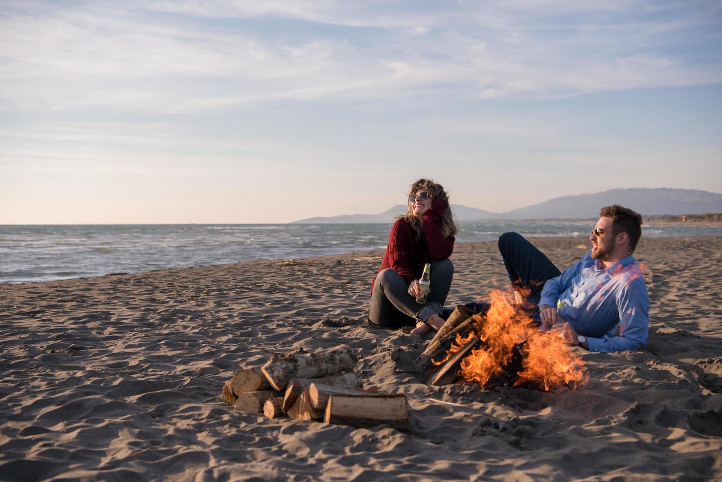 junges Paar sitzt am Strand neben Lagerfeuer und trinkt Bier foto