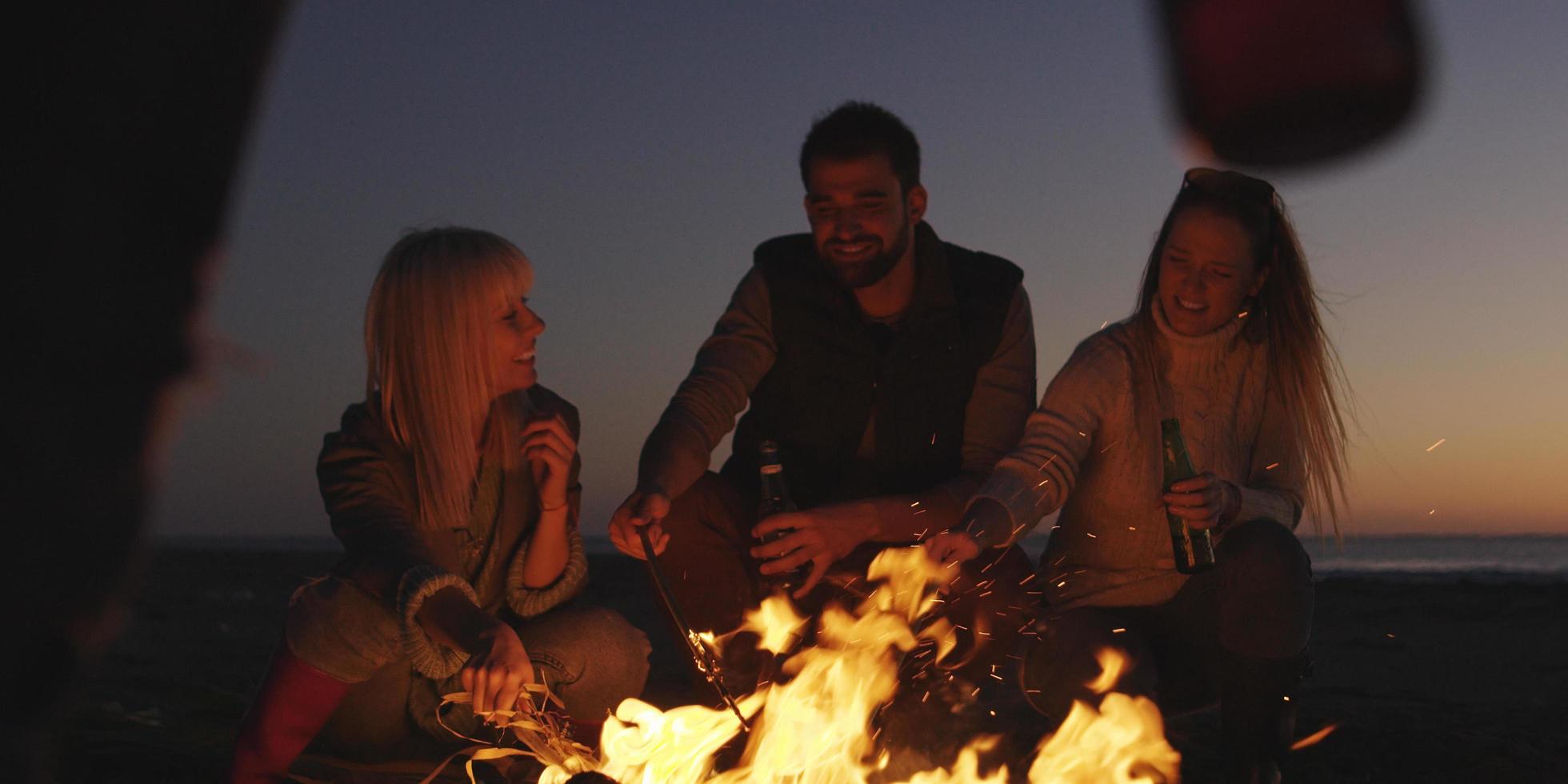 junge freunde, die am lagerfeuer am strand mit bier anstoßen foto