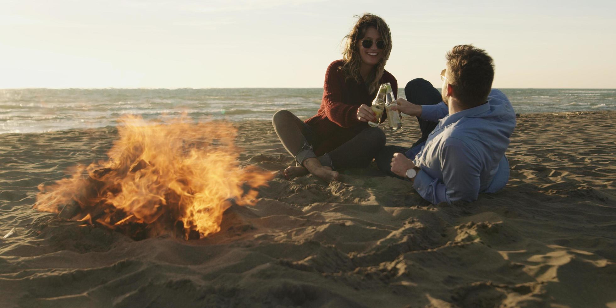 Liebendes junges Paar, das am Strand neben dem Lagerfeuer sitzt und Bier trinkt foto