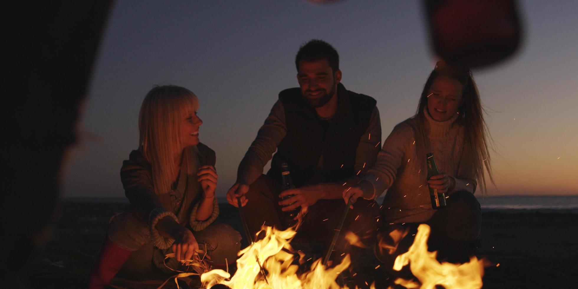 junge freunde, die am lagerfeuer am strand mit bier anstoßen foto