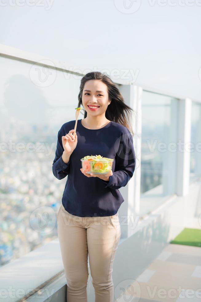 porträt einer attraktiven kaukasischen lächelnden frau, die an einem sonnigen sommertag auf grünem gras im park salat isst, fokus auf gabel mit salat foto