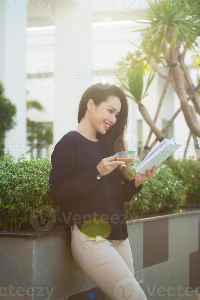 Fröhliche junge Frau, die ein literaturbegeistertes Buch hält, das Romane in der Freizeit auf der Terrasse des Campus-Cafés an sonnigen Tagen analysiert. foto