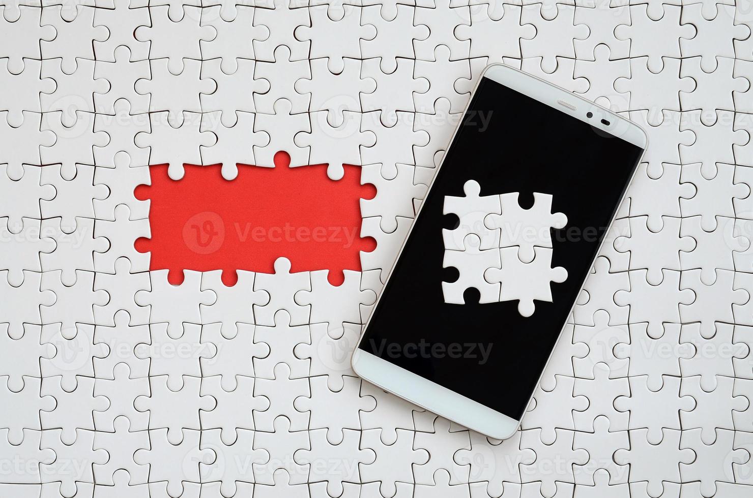 Ein modernes großes Smartphone mit mehreren Puzzleelementen auf dem Touchscreen liegt auf einem weißen Puzzle in zusammengesetztem Zustand mit fehlenden Elementen foto