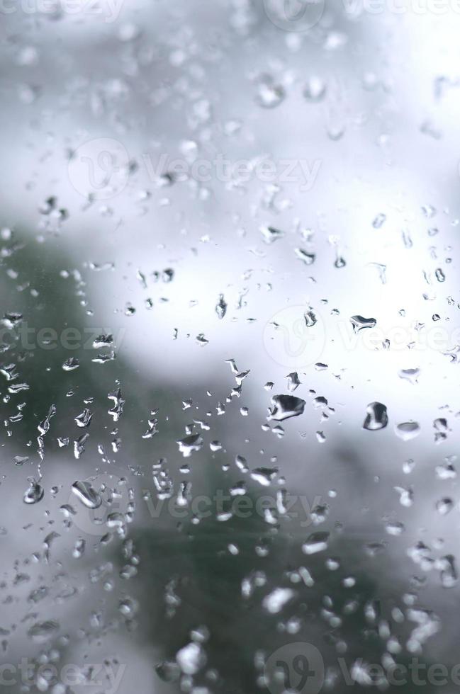 ein Foto von Regentropfen auf dem Fensterglas mit verschwommenem Blick auf die blühenden grünen Bäume. abstraktes Bild, das bewölkte und regnerische Wetterbedingungen zeigt