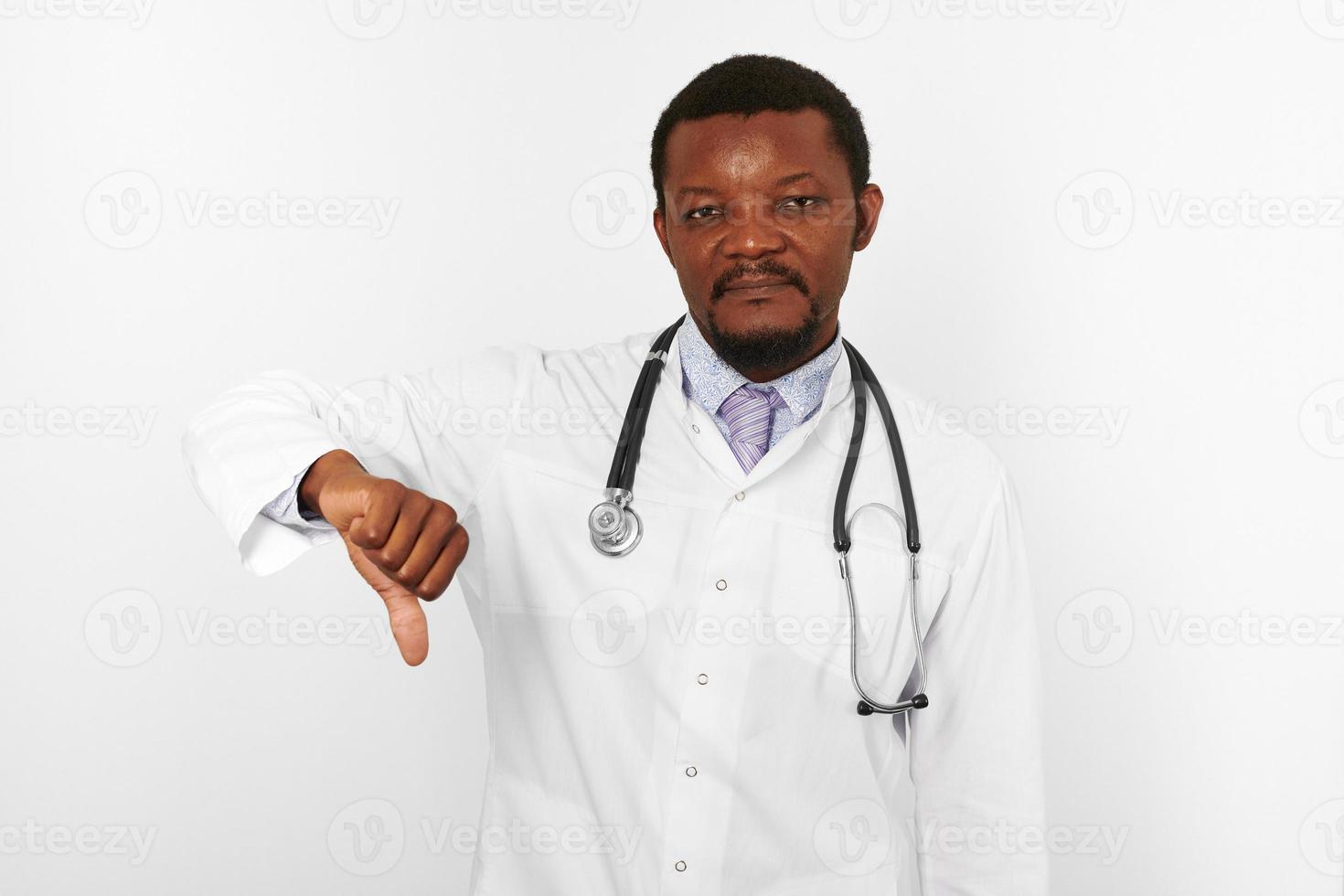 Lächelnder schwarzer bärtiger Arzt in weißer Robe mit Stethoskop zeigt wie Geste, weißer Hintergrund foto