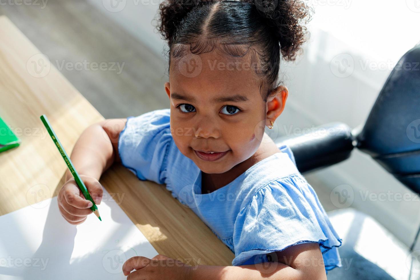 Nahaufnahme eines afroamerikanischen Mädchens, das mit einem Bleistift in der Hand an einem Tisch sitzt, glückliches Lächeln. und schau in die Kamera. foto