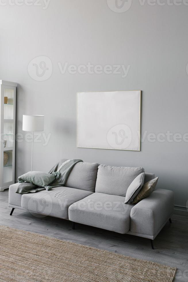 stilvolles minimalistisches Interieur des Wohnzimmers in Grau. sofa mit plaid, stehlampe, beigem teppich und weißem rahmen an der wand foto