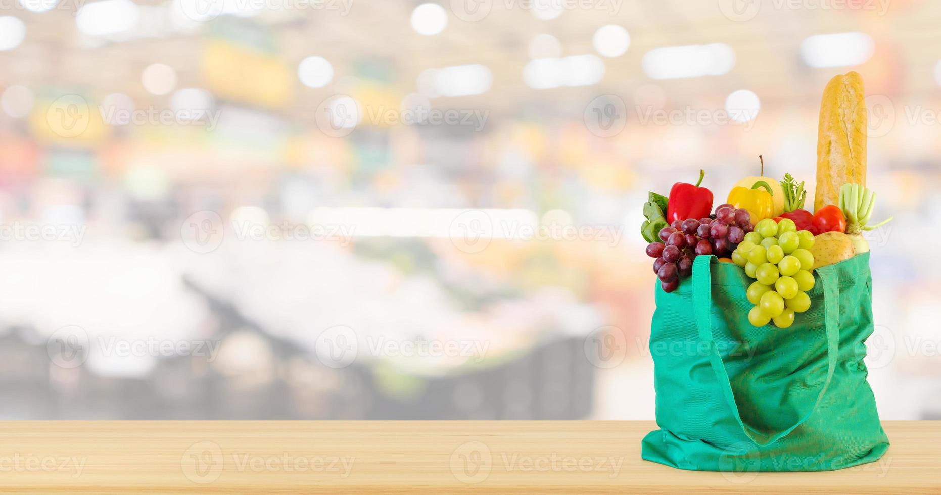 frisches obst und gemüse in wiederverwendbarer grüner einkaufstasche auf holztischplatte mit supermarkt-lebensmittelgeschäft unscharfer defokussierter hintergrund mit bokeh-licht foto