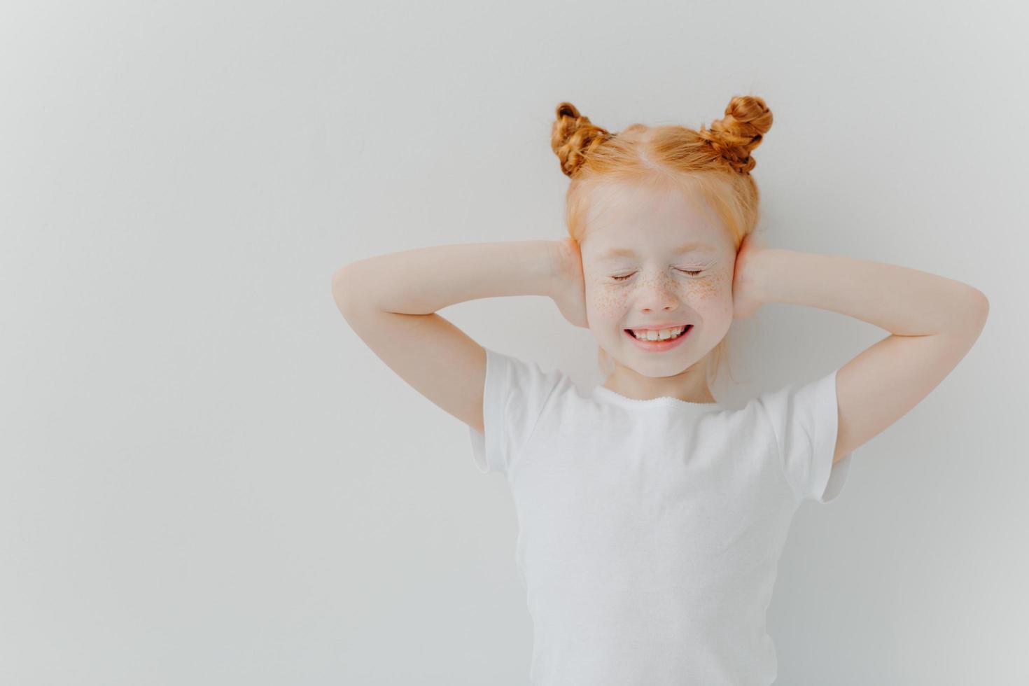 positives, gut aussehendes, rothaariges, schönes mädchen bedeckt die ohren mit den händen, hört sehr laute musik, schließt die augen und lächelt breit, trägt ein weißes t-shirt, steht drinnen, ist auf einer kinderparty foto