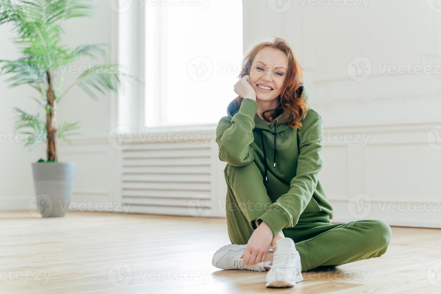 gesundes lebensstilkonzept. schöne schlanke junge frau mit roten welligen haaren, hält die hand unter dem kinn, lächelt glücklich, trägt sportkleidung, hat einen flexiblen körper, sitzt auf dem boden. Frau hat Heimtraining foto