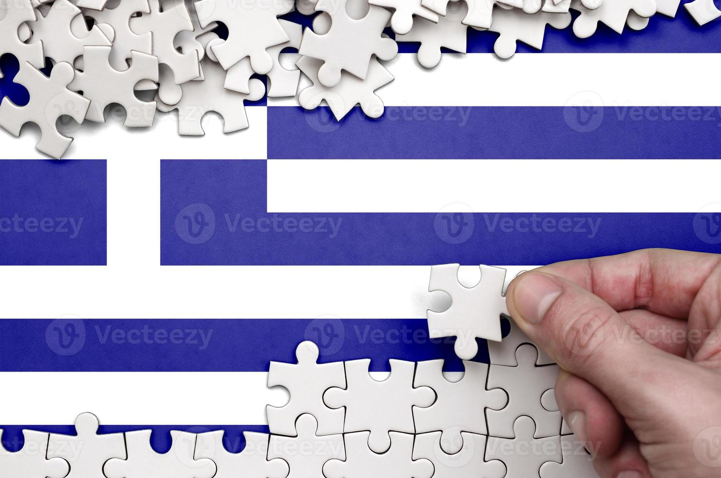 Die griechische Flagge ist auf einem Tisch abgebildet, auf dem die menschliche Hand ein weißes Puzzle faltet foto