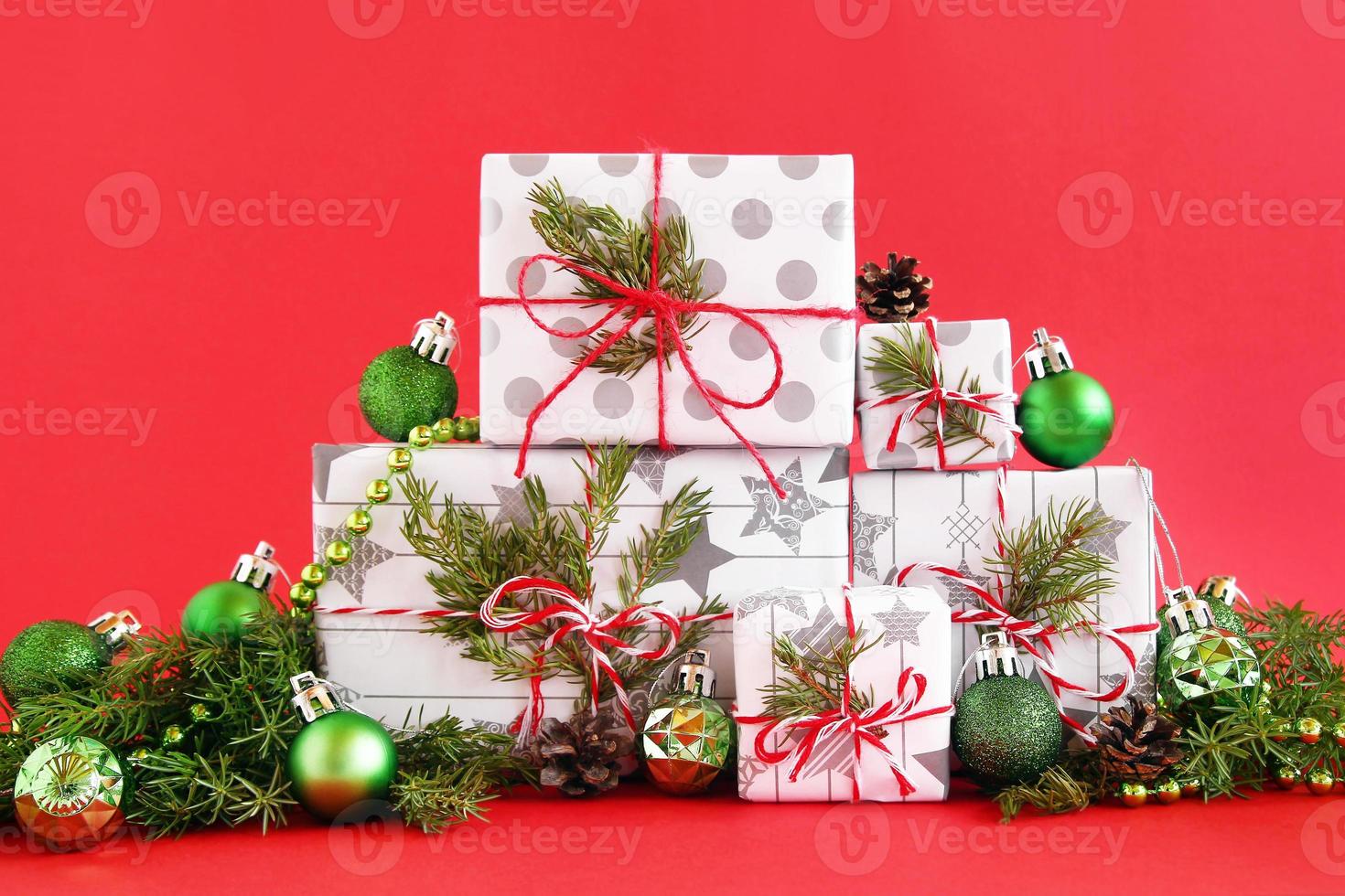 weihnachtsgeschenkboxen aus weiß-grauem papier mit rot-weißen bändern auf rotem hintergrund, verziert mit tannenzweigen, tannenzapfen und glänzend grünem weihnachtsschmuck. foto