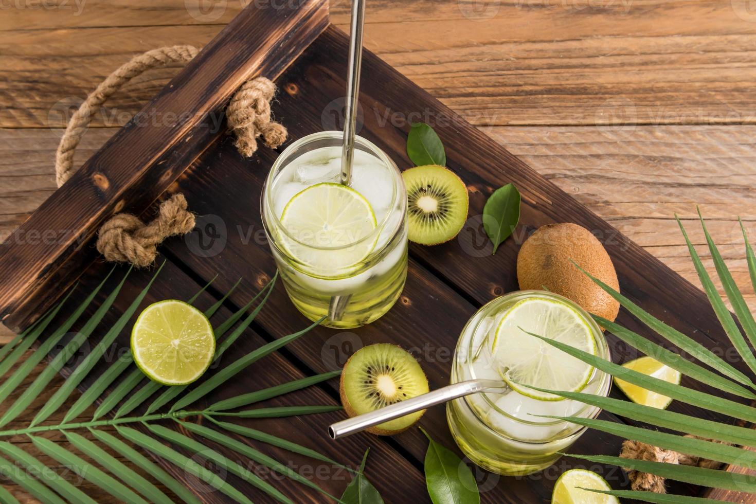 Ein kaltes Erfrischungsgetränk, Vitaminwasser mit Kiwi-Limettenstücken in zwei modernen Gläsern auf einem Holztablett mit tropischen Ästen. Entgiftung. foto