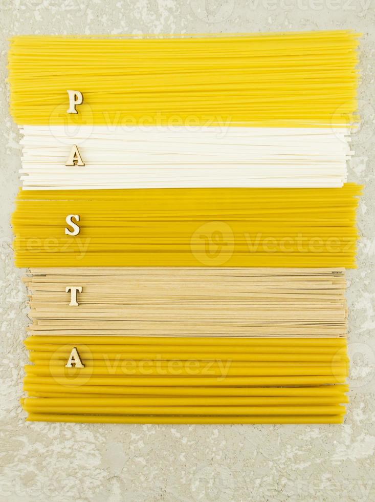 Vertikale flache Draufsicht auf verschiedene italienische Trockenteigwaren auf grauem Hintergrund. Holzbuchstaben mit dem Wort -Pasta. foto