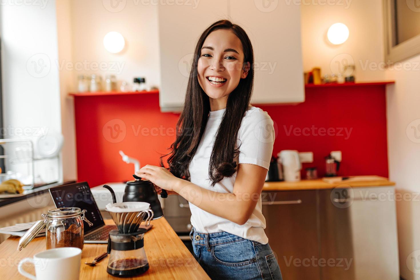 Langhaariges Mädchen lacht aufrichtig und gießt kochendes Wasser in die Kaffeekanne foto
