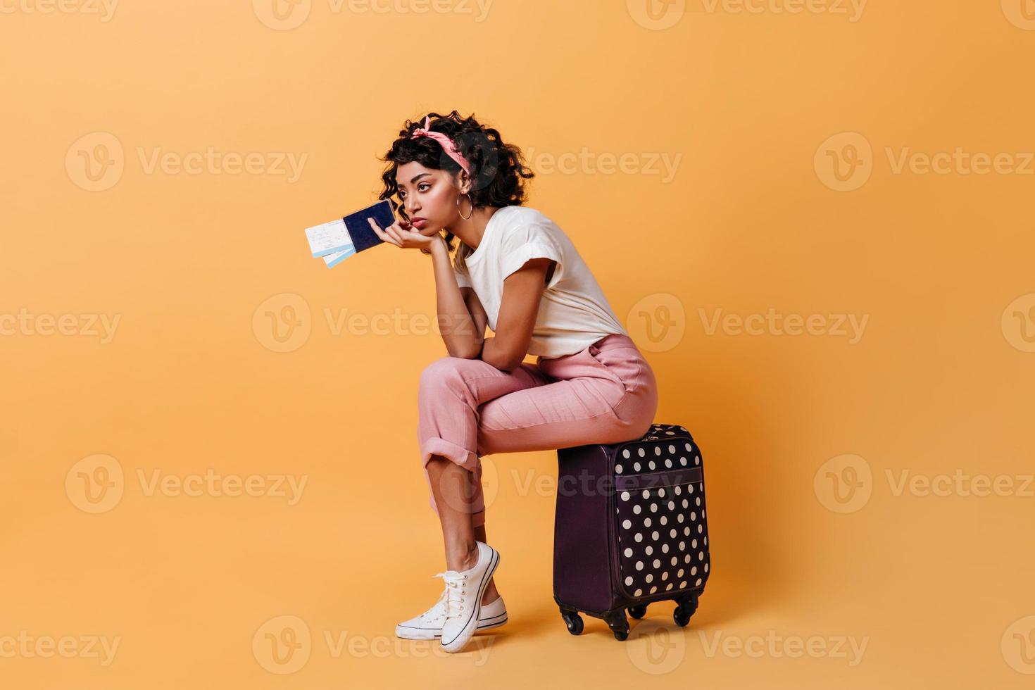 gelangweilte Mischlingsfrau mit Flugtickets. nachdenkliches attraktives mädchen, das auf koffer auf yello sitzt foto