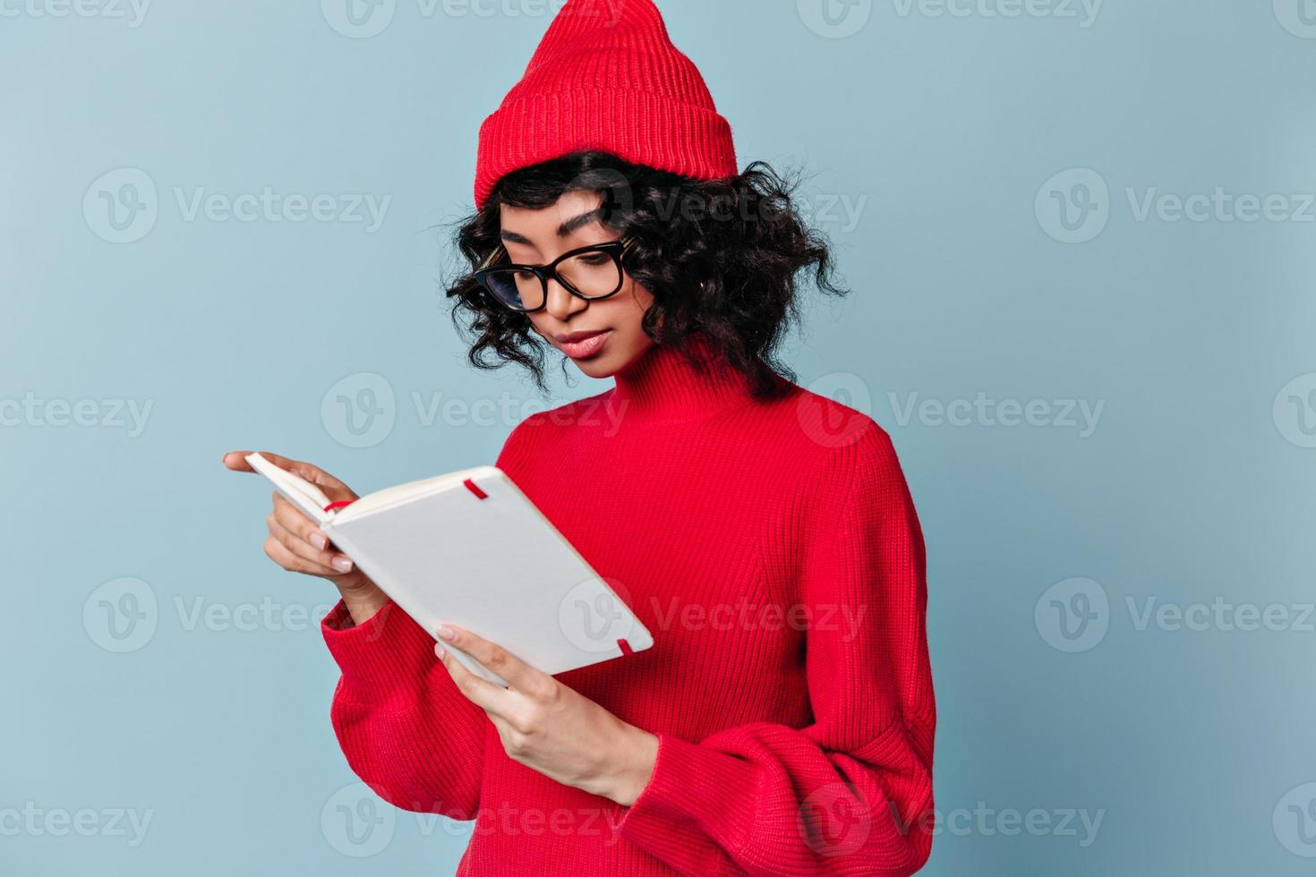 fokussiertes studentenlesenotizbuch. Studioaufnahme eines gemischtrassigen Mädchens mit Brille, das auf Blau steht foto