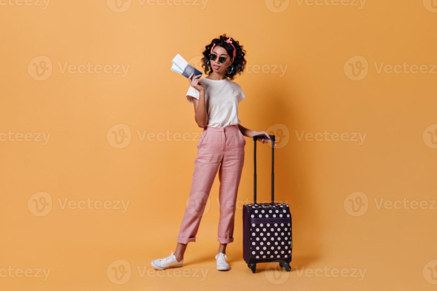 Ganzkörperansicht einer entzückenden Frau mit Koffer und Tickets. weibliche touristin der gemischten rasse, die an steht foto